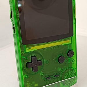 Puede incluir: Una consola de juegos port&aacute;til verde transparente con pantalla negra y botones. La consola tiene un pad direccional, botones A y B, y un bot&oacute;n "SELECT START". El texto "RETROPIXEL" es visible.