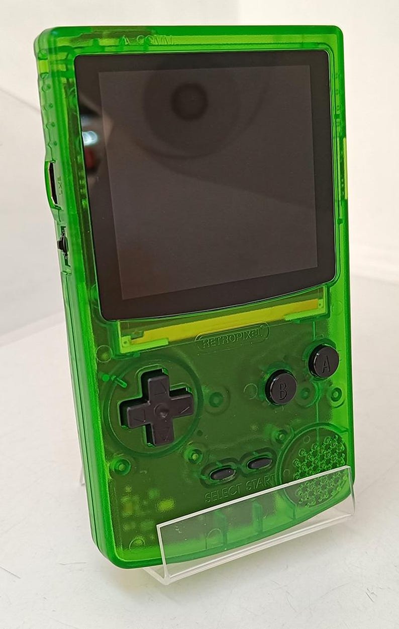 Puede incluir: Una consola de juegos port&aacute;til verde transparente con pantalla negra y botones. La consola tiene un pad direccional, botones A y B, y un bot&oacute;n "SELECT START". El texto "RETROPIXEL" es visible.