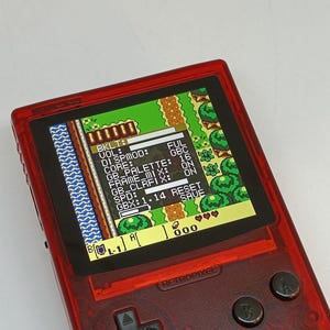 Puede incluir: Una consola de juegos port&aacute;til roja transparente con una pantalla que muestra un videojuego retro. La pantalla muestra una vista desde arriba de un paisaje pixelado con texto y datos del juego. El dispositivo tiene botones y el texto "RETROPIXEL" es visible.