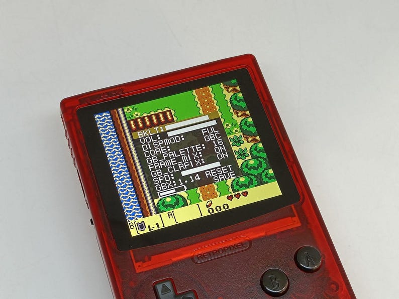 Puede incluir: Una consola de juegos port&aacute;til roja transparente con una pantalla que muestra un videojuego retro. La pantalla muestra una vista desde arriba de un paisaje pixelado con texto y datos del juego. El dispositivo tiene botones y el texto "RETROPIXEL" es visible.