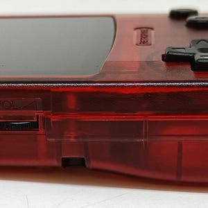 May include: A transparent red handheld gaming console. The device features a black screen, black control buttons, and a USB port. The console's design is visible, showcasing its internal components. The device is designed for gaming.