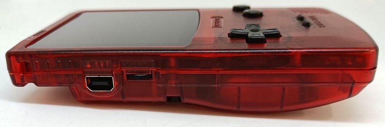 May include: A transparent red handheld gaming console. The device features a black screen, black control buttons, and a USB port. The console's design is visible, showcasing its internal components. The device is designed for gaming.