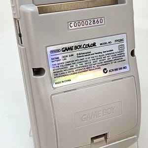 May include: A light gray Game Boy Color handheld game console, viewed from the back. The device has a battery compartment and a label with text including "GAME BOY.COLOR" and "MADE IN CHINA".