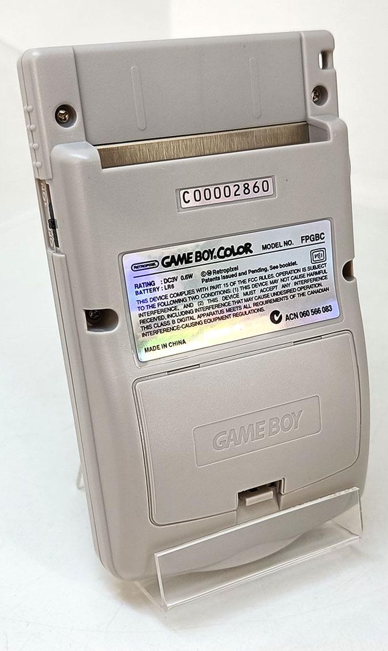 May include: A light gray Game Boy Color handheld game console, viewed from the back. The device has a battery compartment and a label with text including "GAME BOY.COLOR" and "MADE IN CHINA".