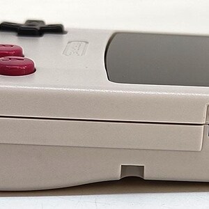 May include: A handheld video game console in a light gray color. It features a black screen, a directional pad, red buttons, and an on/off switch. The console has a retro design.
