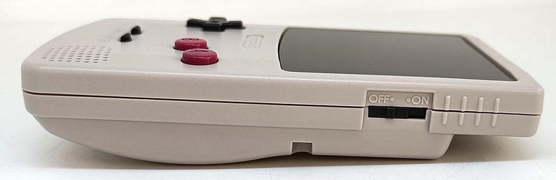 May include: A handheld video game console in a light gray color. It features a black screen, a directional pad, red buttons, and an on/off switch. The console has a retro design.