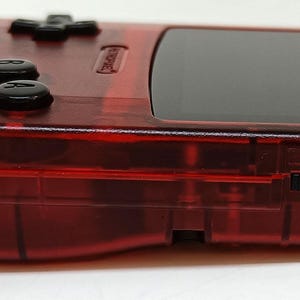 May include: A transparent red handheld gaming console with black buttons and a black screen. The console has a speaker, a directional pad, and two action buttons. The side of the console has a power switch. The device is designed for portable gaming.