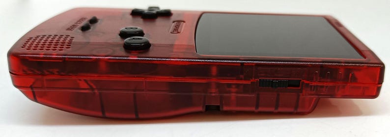 May include: A transparent red handheld gaming console with black buttons and a black screen. The console has a speaker, a directional pad, and two action buttons. The side of the console has a power switch. The device is designed for portable gaming.