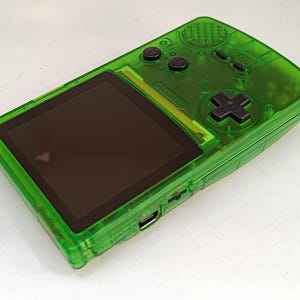 Puede incluir: Una consola de juegos port&aacute;til verde transparente con una pantalla negra y botones negros. La consola tiene un panel direccional y varios botones de funci&oacute;n. El dispositivo es rectangular con bordes redondeados.
