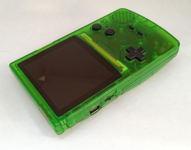 Puede incluir: Una consola de juegos port&aacute;til verde transparente con una pantalla negra y botones negros. La consola tiene un panel direccional y varios botones de funci&oacute;n. El dispositivo es rectangular con bordes redondeados.