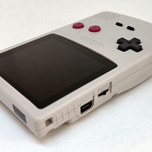 May include: A light gray handheld gaming console with a black screen, red buttons, and a black directional pad. The console has a Nintendo logo and various ports. The design is simple and retro, suggesting a classic gaming device.