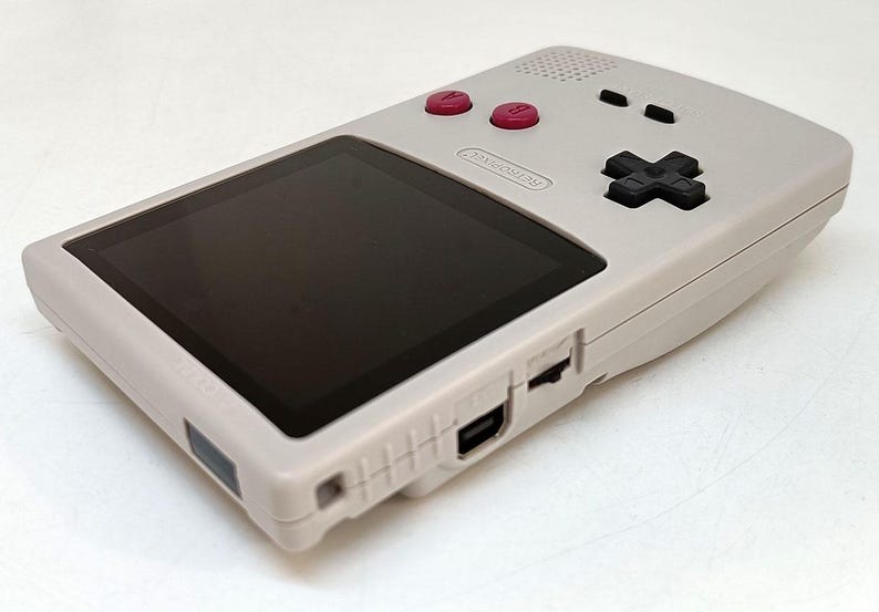 May include: A light gray handheld gaming console with a black screen, red buttons, and a black directional pad. The console has a Nintendo logo and various ports. The design is simple and retro, suggesting a classic gaming device.