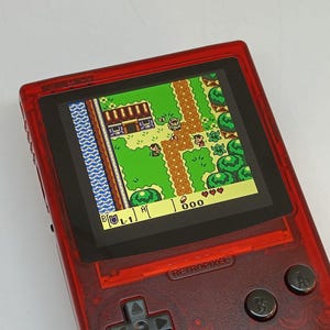 Puede incluir: Una consola de juegos port&aacute;til roja que muestra un videojuego retro con vista superior. La pantalla muestra un paisaje pixelado con personajes, &aacute;rboles y edificios. La consola tiene botones y una cruceta. El texto "RETROPIXEL" es visible.