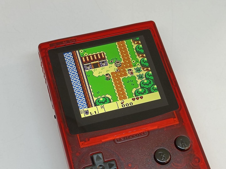 Puede incluir: Una consola de juegos port&aacute;til roja que muestra un videojuego retro con vista superior. La pantalla muestra un paisaje pixelado con personajes, &aacute;rboles y edificios. La consola tiene botones y una cruceta. El texto "RETROPIXEL" es visible.