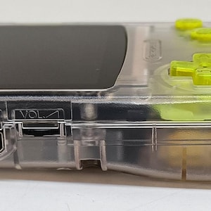 Puede incluir: Una consola de juegos port&aacute;til transparente con botones verde ne&oacute;n y una pantalla negra. La consola tiene una carcasa de pl&aacute;stico transparente, que revela los componentes internos. El mando direccional y los botones son de color verde brillante, contrastando con el cuerpo transparente.