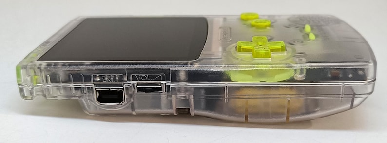 Puede incluir: Una consola de juegos port&aacute;til transparente con botones verde ne&oacute;n y una pantalla negra. La consola tiene una carcasa de pl&aacute;stico transparente, que revela los componentes internos. El mando direccional y los botones son de color verde brillante, contrastando con el cuerpo transparente.