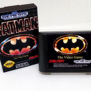 May include: A black Sega Genesis game cartridge for Batman: The Video Game. The cartridge features a yellow and orange Batman logo on a black background. The text "SUNSOFT" and "The Video Game" are printed on the cartridge.