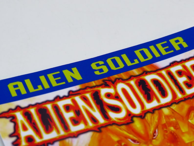 Może przedstawiać: Niebiesko-zielony baner z napisem "ALIEN SOLDIER" białymi literami. Baner znajduje się nad ognistym pomarańczowo-ż&oacute;łtym tłem z napisem "ALIEN SOLDIER" białymi literami.