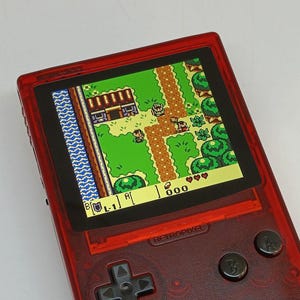 Puede incluir: Una consola de juegos port&aacute;til roja que muestra un videojuego retro con vista superior. La pantalla muestra un paisaje verde con personajes y edificios pixelados. La consola tiene una cruceta, botones y el texto "RETROPIXEL".