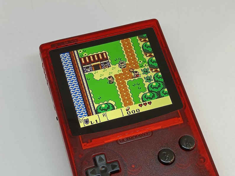 Puede incluir: Una consola de juegos port&aacute;til roja que muestra un videojuego retro con vista superior. La pantalla muestra un paisaje verde con personajes y edificios pixelados. La consola tiene una cruceta, botones y el texto "RETROPIXEL".