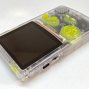 Puede incluir: Una consola de juegos port&aacute;til transparente con botones verde ne&oacute;n y una pantalla negra. La consola tiene una carcasa de pl&aacute;stico transparente, con un pad direccional y botones en verde brillante. La pantalla es rectangular y negra.