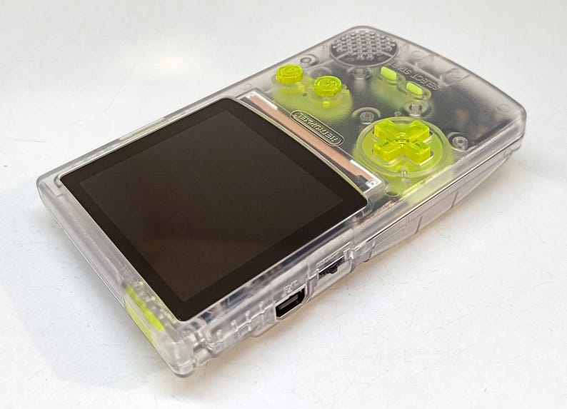 Puede incluir: Una consola de juegos port&aacute;til transparente con botones verde ne&oacute;n y una pantalla negra. La consola tiene una carcasa de pl&aacute;stico transparente, con un pad direccional y botones en verde brillante. La pantalla es rectangular y negra.