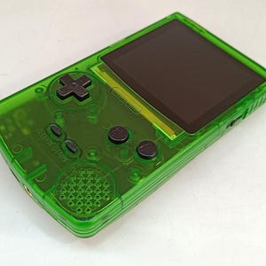 Puede incluir: Consola de juegos port&aacute;til verde transparente con pantalla negra y botones negros. La consola cuenta con una cruceta, botones select y start, y botones A y B. El logotipo de Nintendo es visible.