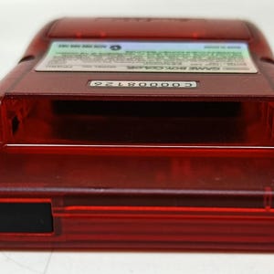 May include: A transparent red Game Boy Color handheld console. The device has a rectangular shape with rounded edges and a black rectangular power button. The top of the device has a label with text and a serial number.