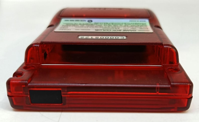 May include: A transparent red Game Boy Color handheld console. The device has a rectangular shape with rounded edges and a black rectangular power button. The top of the device has a label with text and a serial number.