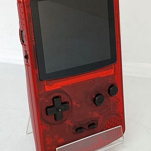 May include: A transparent red handheld gaming console with a black screen and black buttons. The console has a directional pad, A and B buttons, and the text "RETROPIXEL". It is displayed on a clear stand.