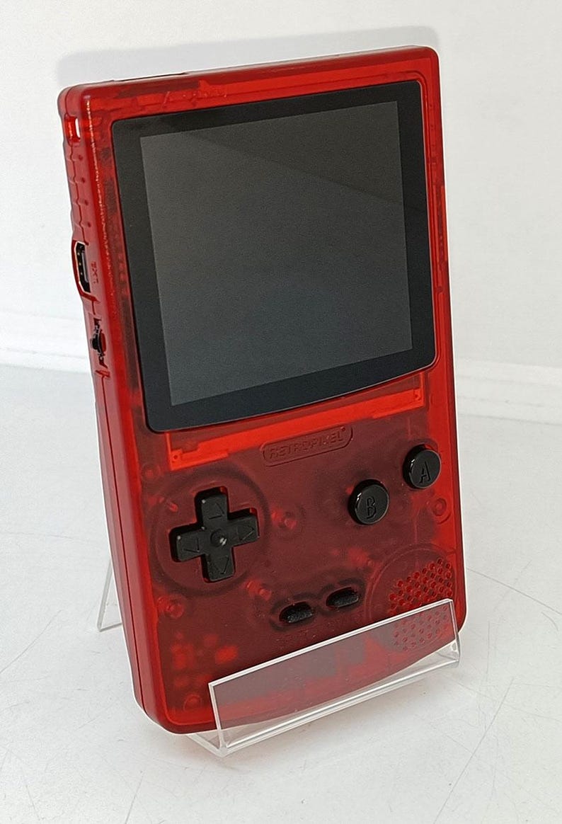 May include: A transparent red handheld gaming console with a black screen and black buttons. The console has a directional pad, A and B buttons, and the text "RETROPIXEL". It is displayed on a clear stand.