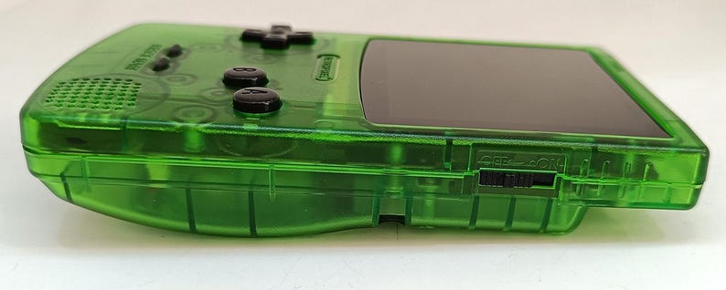 Puede incluir: Una consola de juegos port&aacute;til verde transparente. El dispositivo presenta una pantalla negra, botones de control negros y un altavoz. El dise&ntilde;o de la consola incluye una cruceta, botones de acci&oacute;n y un interruptor de encendido. La carcasa de pl&aacute;stico es de un verde vibrante y transparente.
