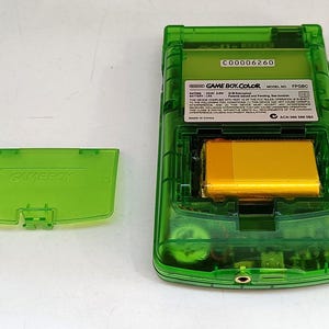 Puede incluir: Una consola de juegos port&aacute;til Game Boy Color verde transparente, con el compartimento de la bater&iacute;a abierto. La bater&iacute;a es de color dorado. La tapa de la bater&iacute;a est&aacute; separada, mostrando el logotipo de Game Boy. La parte trasera de la consola tiene una etiqueta con texto.