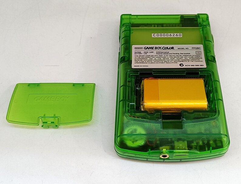 Puede incluir: Una consola de juegos port&aacute;til Game Boy Color verde transparente, con el compartimento de la bater&iacute;a abierto. La bater&iacute;a es de color dorado. La tapa de la bater&iacute;a est&aacute; separada, mostrando el logotipo de Game Boy. La parte trasera de la consola tiene una etiqueta con texto.