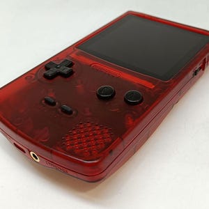 May include: A transparent red handheld gaming console with a black screen, directional pad, and buttons. The console has a speaker, headphone jack, and a power switch. The Nintendo logo is visible.