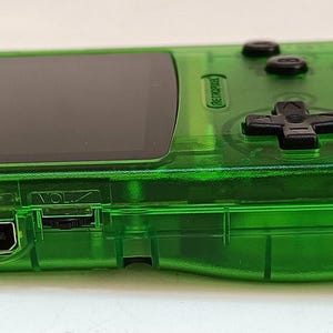 Puede incluir: Una consola de juegos port&aacute;til verde transparente con una pantalla negra y botones negros. La consola tiene forma rectangular con bordes redondeados y una cruceta. Tiene un puerto USB y control de volumen.