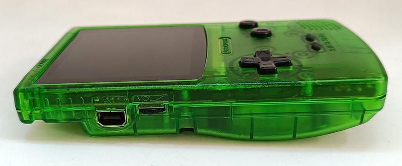 Puede incluir: Una consola de juegos port&aacute;til verde transparente con una pantalla negra y botones negros. La consola tiene forma rectangular con bordes redondeados y una cruceta. Tiene un puerto USB y control de volumen.