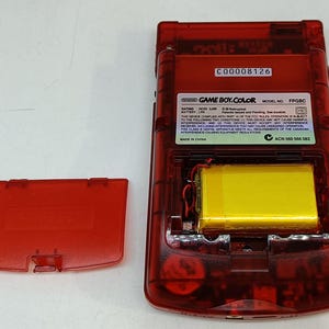 May include: A transparent red Game Boy Color handheld game console, with the battery compartment open. The yellow battery is visible inside. The battery cover is detached and lying to the side. The text "GAME BOY.COLOR" is visible.