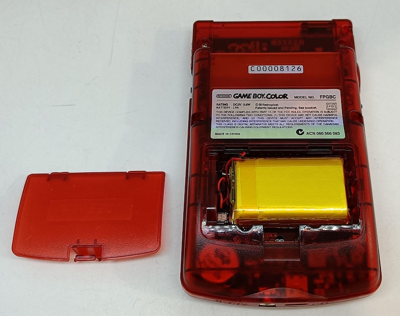May include: A transparent red Game Boy Color handheld game console, with the battery compartment open. The yellow battery is visible inside. The battery cover is detached and lying to the side. The text "GAME BOY.COLOR" is visible.