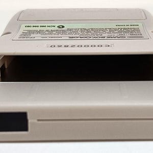 May include: A light gray Game Boy Color handheld game console. The device has a rectangular shape with a large opening for the game cartridge. The back of the device has a label with text. The console is made in China.