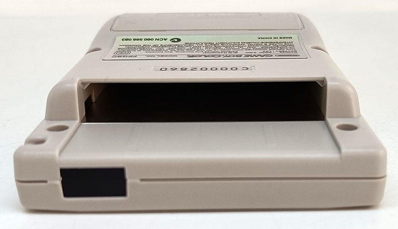 May include: A light gray Game Boy Color handheld game console. The device has a rectangular shape with a large opening for the game cartridge. The back of the device has a label with text. The console is made in China.