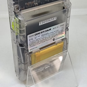 Puede incluir: Una consola de juegos port&aacute;til Game Boy Color transparente. El dispositivo tiene una carcasa de pl&aacute;stico transparente, que revela los componentes internos. La etiqueta dice "GAME BOY COLOR" y "MADE IN CHINA". El n&uacute;mero de modelo es FPGBC.