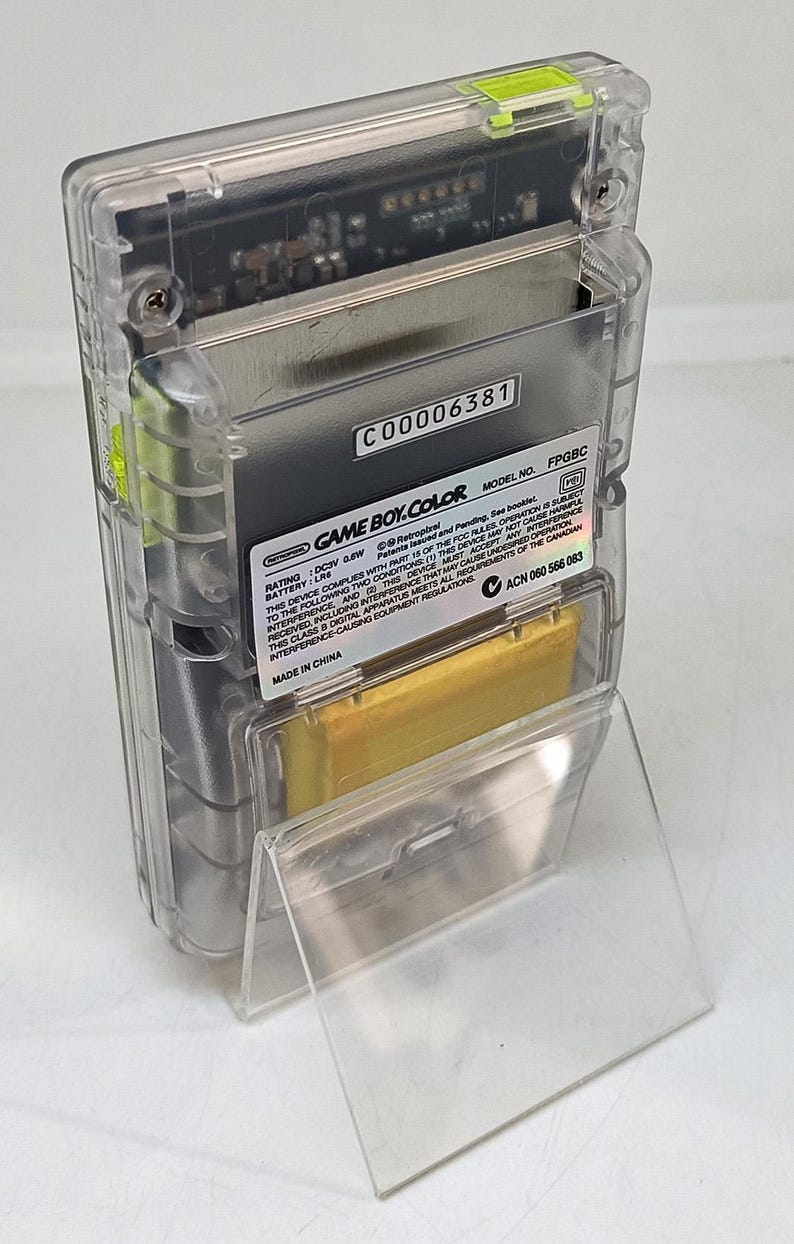 Puede incluir: Una consola de juegos port&aacute;til Game Boy Color transparente. El dispositivo tiene una carcasa de pl&aacute;stico transparente, que revela los componentes internos. La etiqueta dice "GAME BOY COLOR" y "MADE IN CHINA". El n&uacute;mero de modelo es FPGBC.