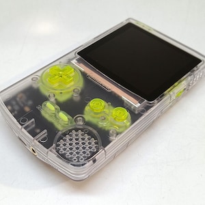 Puede incluir: Una consola de juegos port&aacute;til transparente con botones verde ne&oacute;n y una pantalla negra. La carcasa transparente revela los componentes internos. La cruceta y los botones son de color verde brillante, contrastando con la pantalla negra.