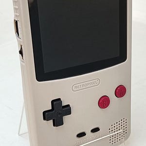 Peut inclure: Console de jeu portable gris clair avec un écran noir. Elle comporte des boutons A et B rouges, une croix directionnelle et les textes "RETROPIXEL" et "SELECT START". La console est posée sur un support transparent.