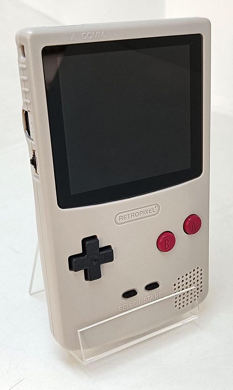 May include: A light gray handheld gaming console with a black screen. It features red A and B buttons, a directional pad, and the text "RETROPIXEL" and "SELECT START". The console is propped up on a clear stand.