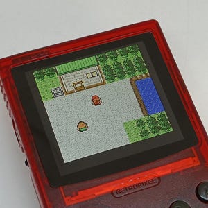 Puede incluir: Una consola de juegos port&aacute;til roja que muestra una escena de videojuego retro pixelada. La pantalla muestra una vista desde arriba de un edificio, &aacute;rboles y dos personajes. La consola tiene la palabra "RETROPIXEL" impresa en la parte inferior.