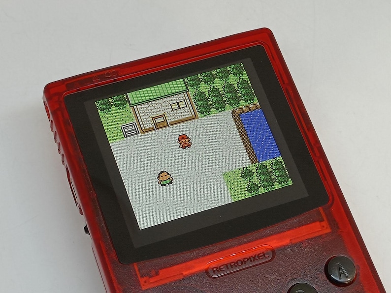 Puede incluir: Una consola de juegos port&aacute;til roja que muestra una escena de videojuego retro pixelada. La pantalla muestra una vista desde arriba de un edificio, &aacute;rboles y dos personajes. La consola tiene la palabra "RETROPIXEL" impresa en la parte inferior.