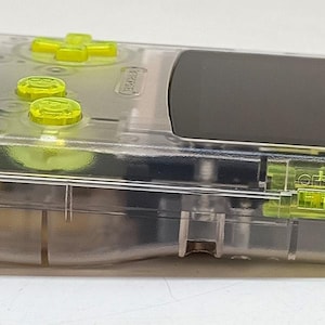 Puede incluir: Una consola de juegos port&aacute;til transparente con botones y detalles en amarillo ne&oacute;n. El dispositivo tiene una pantalla negra y una carcasa de pl&aacute;stico transparente, que revela los componentes internos. El logotipo de Nintendo es visible.