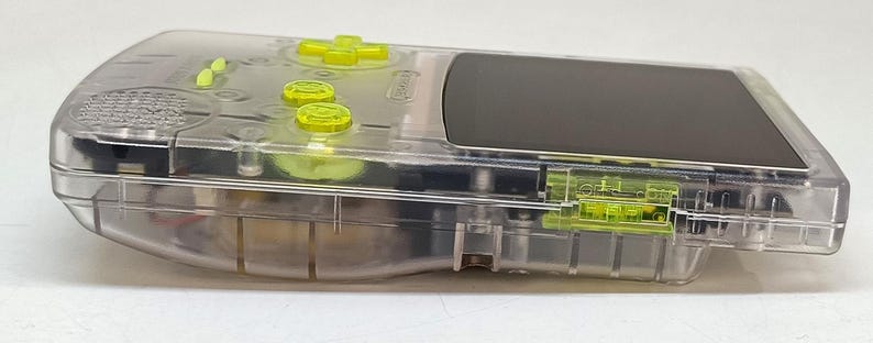 Puede incluir: Una consola de juegos port&aacute;til transparente con botones y detalles en amarillo ne&oacute;n. El dispositivo tiene una pantalla negra y una carcasa de pl&aacute;stico transparente, que revela los componentes internos. El logotipo de Nintendo es visible.